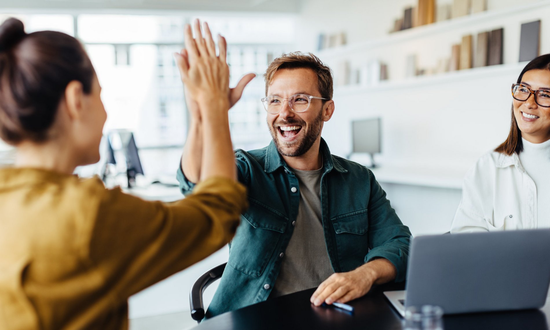 Zwei lächelnde Personen in einem Besprechungsraum geben sich ein High-Five. Eine dritte Person beobachtet die Szene. Die Atmosphäre ist freundlich und positiv, was auf eine erfolgreiche Zusammenarbeit hindeutet. Ein Laptop und Büroeinrichtungen sind im Hintergrund sichtbar.