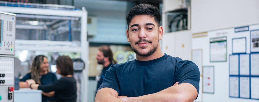 Ein Mann steht vor einer Maschine und schaut direkt in die Kamera, lächelnd und freundlich.