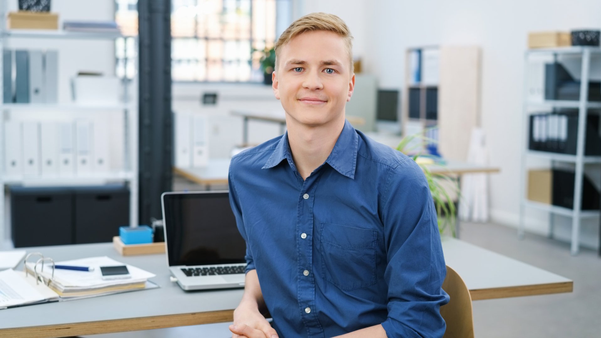 Ein junger Mann mit blonden Haaren sitzt in einem modernen Büro. Er trägt ein blaues Hemd und lächelt freundlich. Auf dem Tisch vor ihm liegen Unterlagen und ein Laptop. Der Hintergrund zeigt Regale mit Aktenordnern.