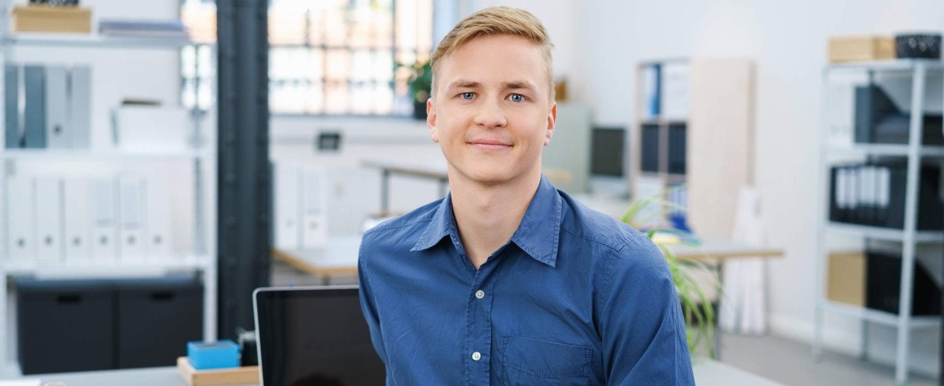 Ein junger Mann mit blonden Haaren sitzt in einem modernen Büro. Er trägt ein blaues Hemd und lächelt freundlich. Auf dem Tisch vor ihm liegen Unterlagen und ein Laptop. Der Hintergrund zeigt Regale mit Aktenordnern.