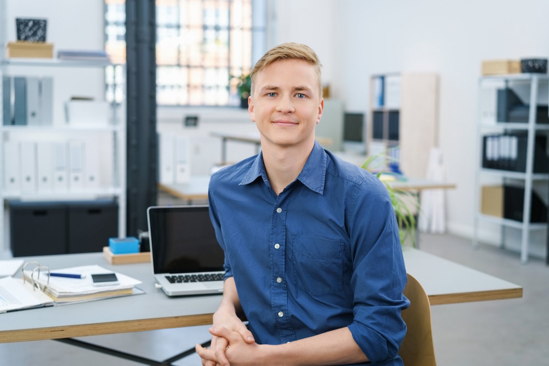 Ein junger Mann mit blonden Haaren sitzt in einem modernen Büro. Er trägt ein blaues Hemd und lächelt freundlich. Auf dem Tisch vor ihm liegen Unterlagen und ein Laptop. Der Hintergrund zeigt Regale mit Aktenordnern.