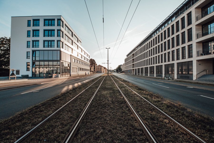 Eine symmetrische Straßenansicht mit Straßenbahnschienen in der Mitte, die auf beiden Seiten von modernen, eckigen Gebäuden gesäumt sind.
