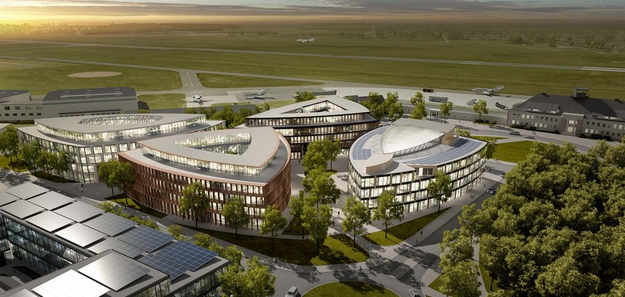 Luftaufnahme eines modernen Architekturkomplexes mit mehreren einzigartig geformten Gebäuden, umgeben von Grün. Der Komplex liegt neben einer Landebahn, auf der kleine Flugzeuge zu sehen sind, und am Horizont ist bei Sonnenaufgang oder Sonnenuntergang ein bewölkter Himmel zu sehen.
