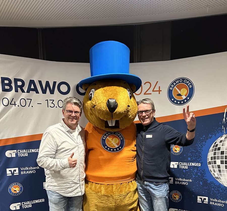 Drei Personen posieren für ein Foto: Zwei Erwachsene stehen auf beiden Seiten einer Person in einem großen Maskottchenkostüm. Das Maskottchen ist ein gelbes Tier, das ein orangefarbenes Hemd und einen blauen Zylinder trägt. Die Erwachsenen und das Maskottchen lächeln vor einem Banner mit Veranstaltungslogos und -daten.