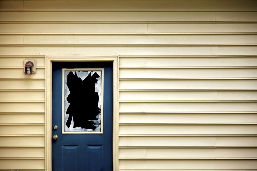 Eine blaue Haustür mit einer beschädigten Fensterscheibe, die mit schwarzem Material verklebt ist. Die Tür befindet sich an einer gelben Wand. Ein Türknopf und ein kleines Licht sind sichtbar. Die Aufmachung vermittelt einen unsicheren Eindruck.