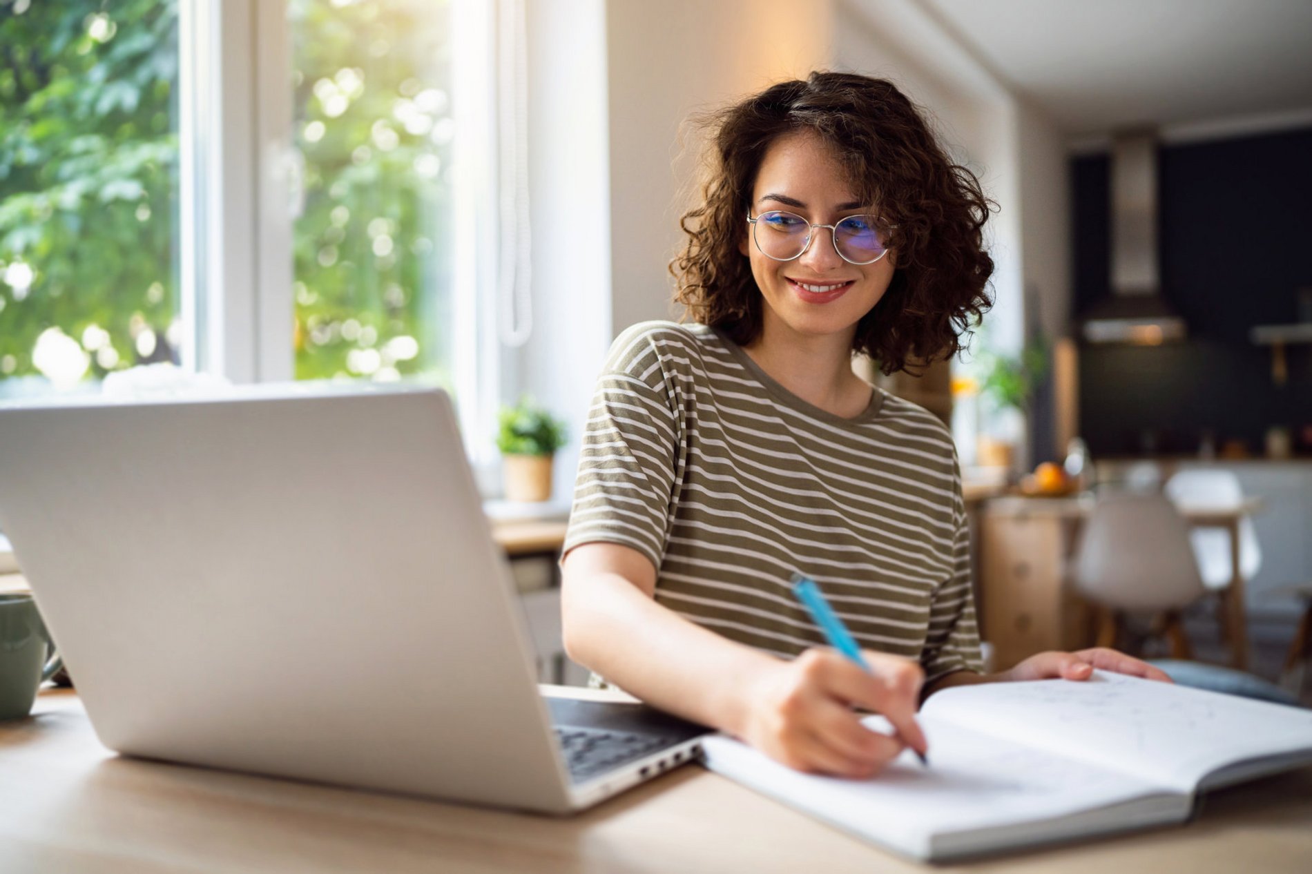 Eine lächelnde junge Frau mit lockigem Haar sitzt an einem Tisch und schreibt in ein Notizbuch, während ein Laptop vor ihr steht. Im Hintergrund sind Pflanzen und ein heller Raum zu sehen, der ein freundliches Arbeitsumfeld schafft.