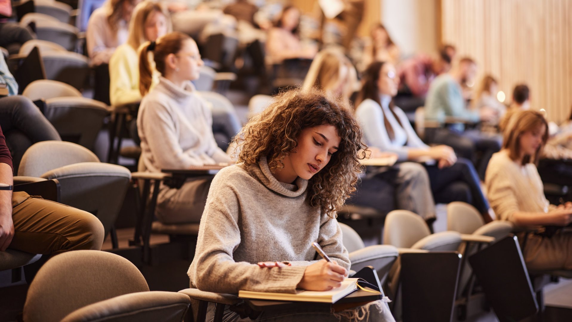 Eine Studentin sitzt in einem Hörsaal und macht sich Notizen in ein Notizbuch. Im Hintergrund sind weitere Studierende zu sehen, die aufmerksam zuhören. Der Raum hat eine moderne Holzverkleidung und ist mit Sitzreihen ausgestattet.