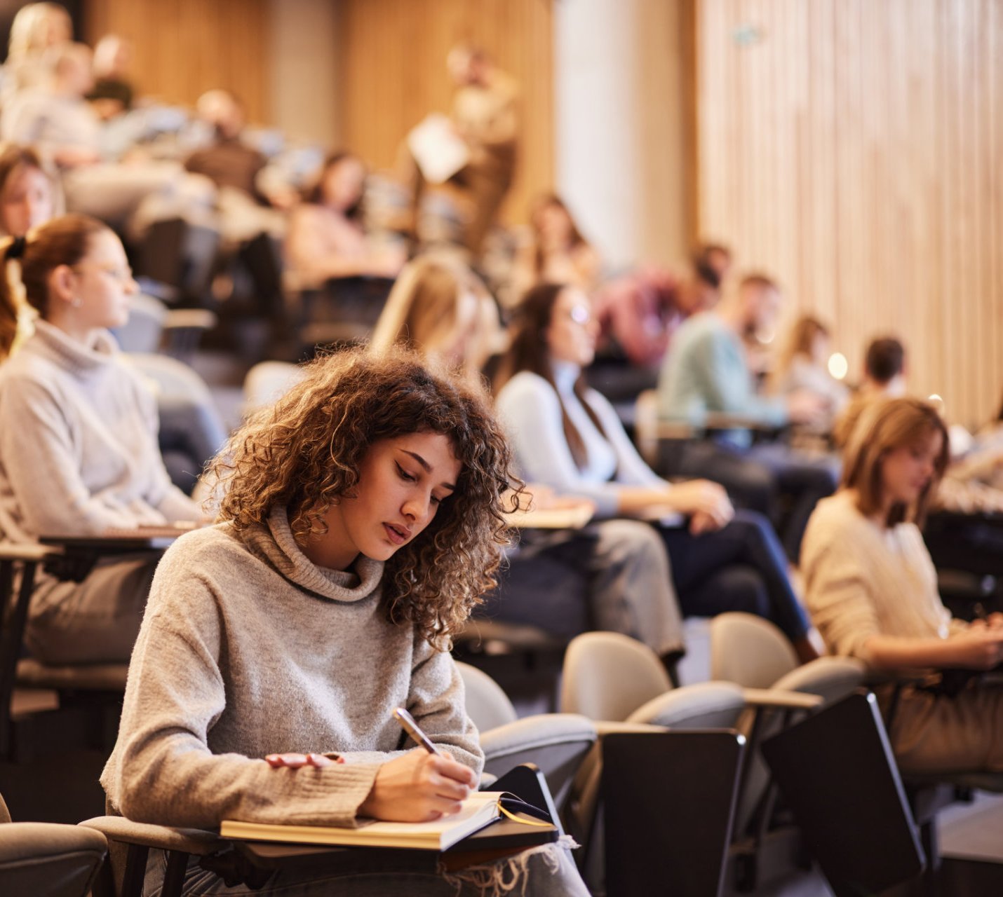 Eine Studentin sitzt in einem Hörsaal und macht sich Notizen in ein Notizbuch. Im Hintergrund sind weitere Studierende zu sehen, die aufmerksam zuhören. Der Raum hat eine moderne Holzverkleidung und ist mit Sitzreihen ausgestattet.