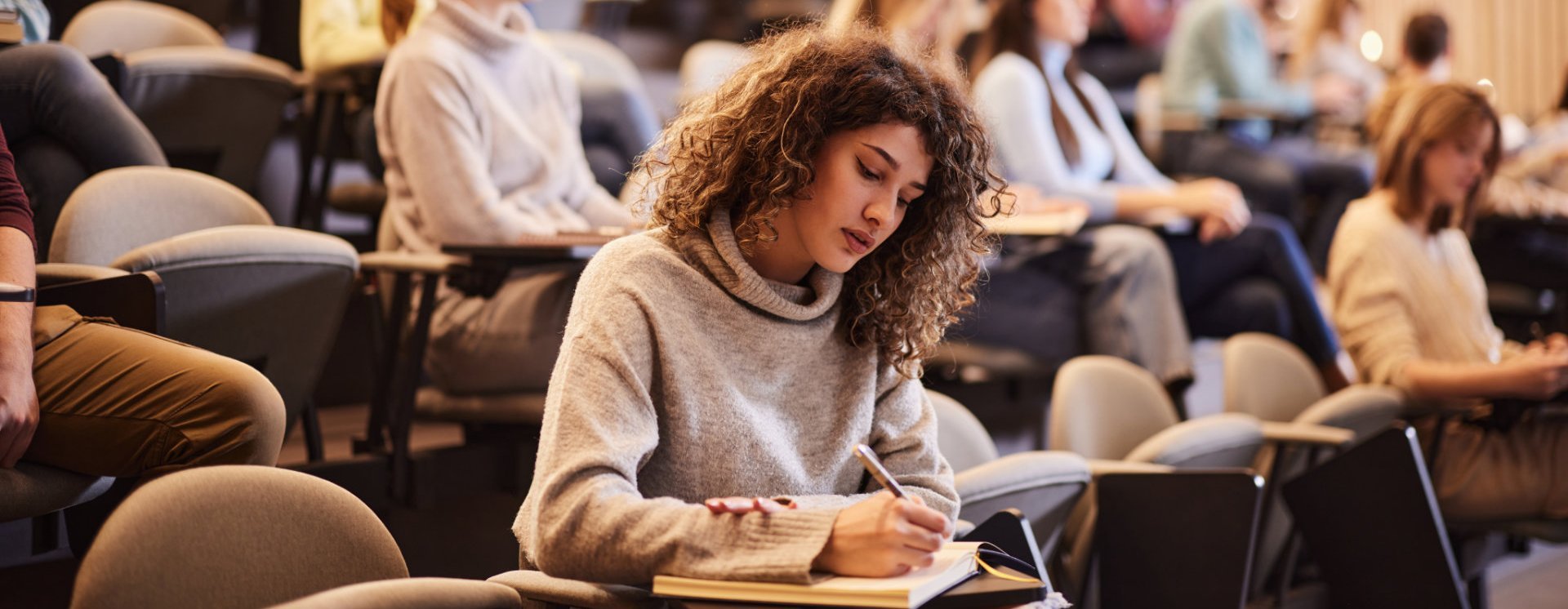 Eine Studentin sitzt in einem Hörsaal und macht sich Notizen in ein Notizbuch. Im Hintergrund sind weitere Studierende zu sehen, die aufmerksam zuhören. Der Raum hat eine moderne Holzverkleidung und ist mit Sitzreihen ausgestattet.