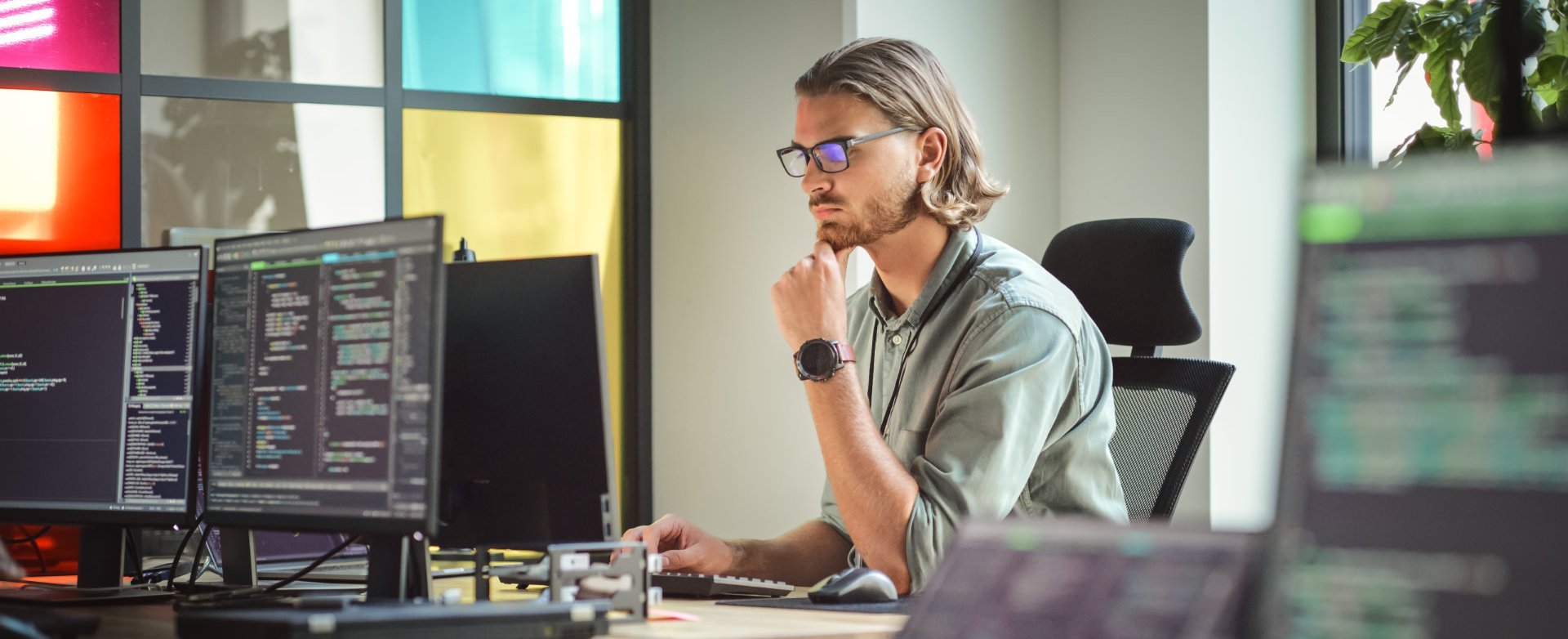 Ein junger Mann mit Brille sitzt an einem Schreibtisch vor mehreren Computermonitoren, auf denen Code zu sehen ist. Er schaut konzentriert auf den Bildschirm und scheint an einer Programmieraufgabe zu arbeiten. Im Hintergrund sind bunte Fenster und Pflanzen sichtbar.