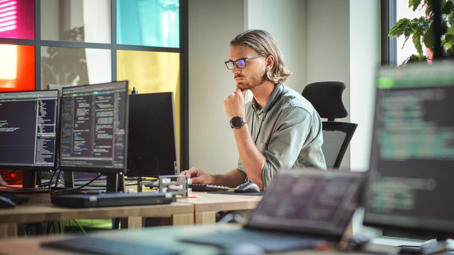 Ein junger Mann mit Brille sitzt an einem Schreibtisch vor mehreren Computermonitoren, auf denen Code zu sehen ist. Er schaut konzentriert auf den Bildschirm und scheint an einer Programmieraufgabe zu arbeiten. Im Hintergrund sind bunte Fenster und Pflanzen sichtbar.