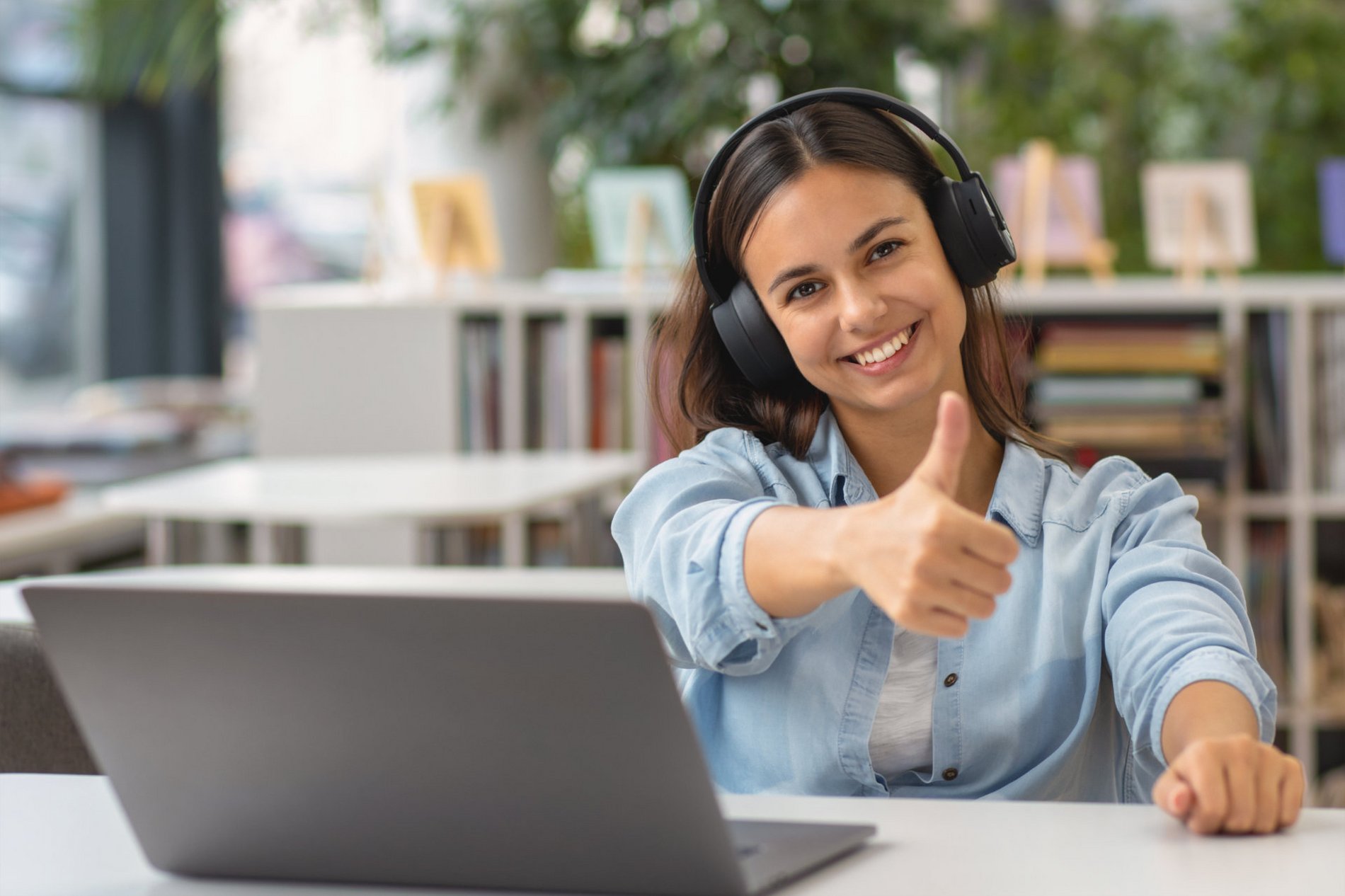 Eine lächelnde Frau mit Kopfhörern sitzt an einem Tisch und gibt mit ihrem Daumen ein positives Zeichen. Vor ihr steht ein Laptop. Im Hintergrund sind Bücherregale und eine freundliche, helle Umgebung zu sehen.