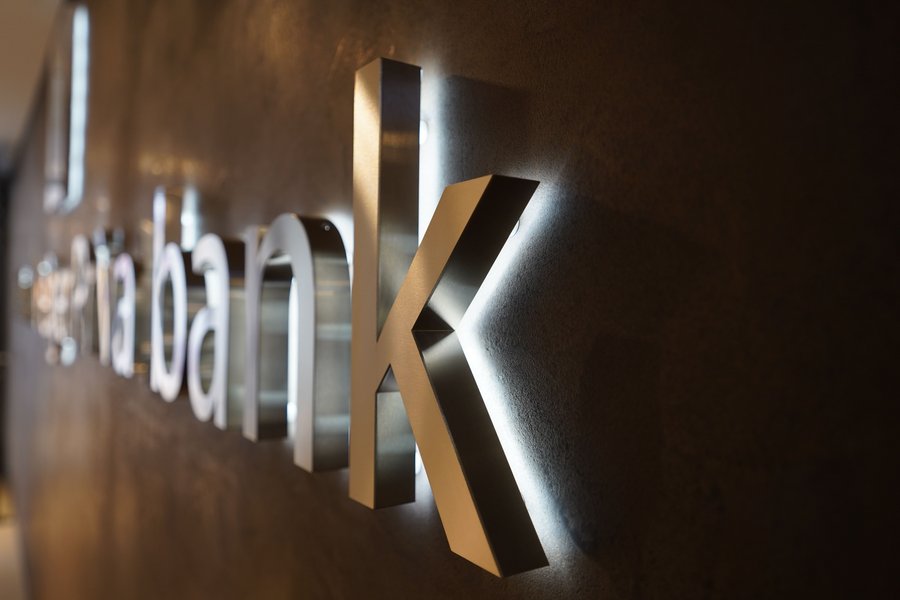 Eine Nahaufnahme eines eleganten, metallischen Banklogos, das an einer Wand angebracht ist. Eine sanfte Hintergrundbeleuchtung hebt die dreidimensionalen Buchstaben hervor. Das Wort „Bank“ ist deutlich zu erkennen und weist ein stilvolles und modernes Design auf, das sich von der strukturierten Wand abhebt.