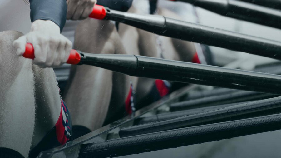 Nahaufnahme von Ruderern in einem Boot, die jeweils gleichzeitig die Ruder mit roten Griffen umklammern.