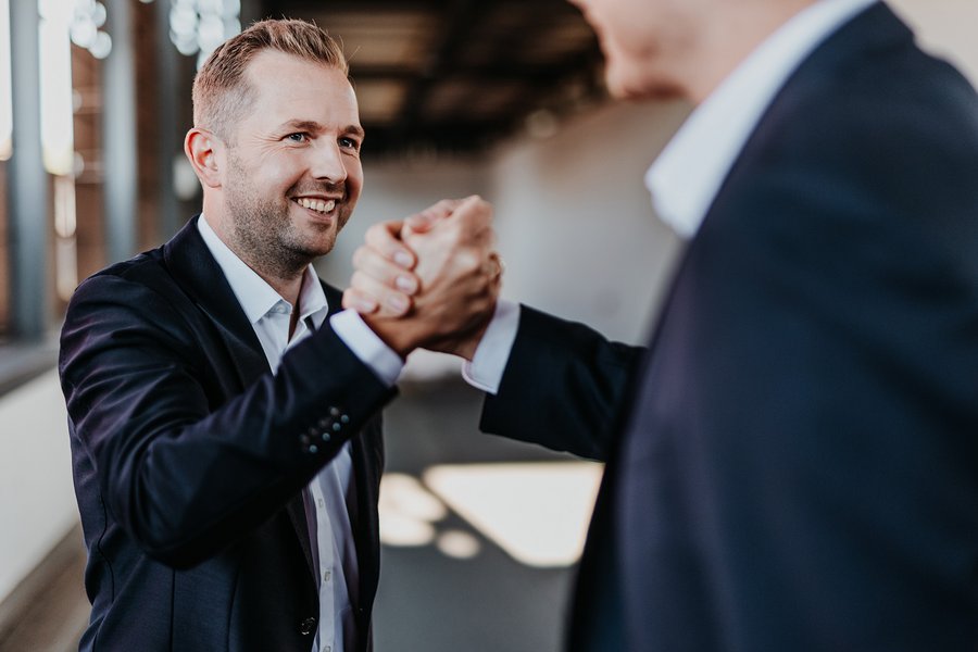 Ein Mann im Anzug schüttelt einer anderen Person im Vordergrund, die nicht im Fokus steht, begeistert die Hand. Beide tragen dunkle Geschäftskleidung in einem gut beleuchteten, modernen Büroraum. Der lächelnde Mann wirkt freundlich und engagiert.